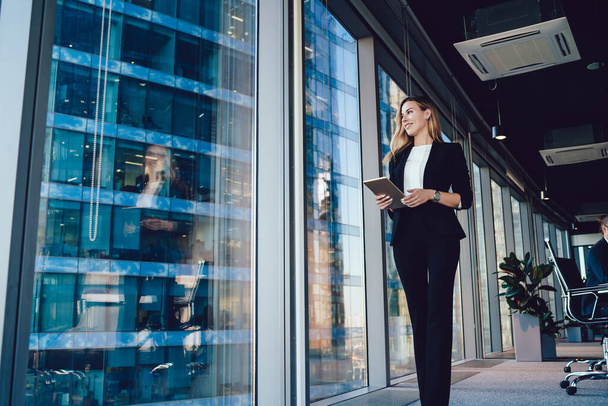 Caucasian female entrepreneur dressed in formal wear going near panoramic window in office, confident business woman with modern digital tablet looking away during working day in enterprise - Foto, Imagen