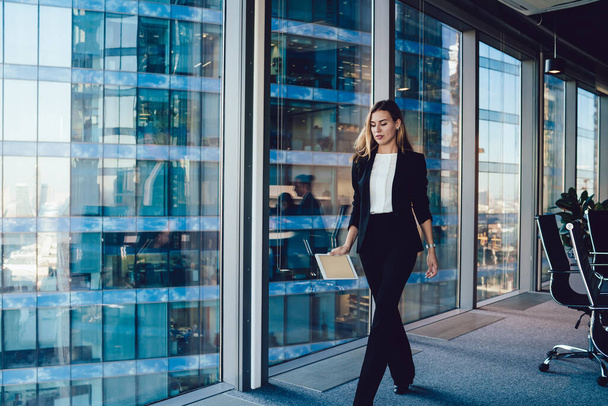 Confident female proud ceo of corporation dressed in formal wear passing glass panoramic window in office interior with mockup modern tablet, intelligent business woman going to company meeting - Photo, image