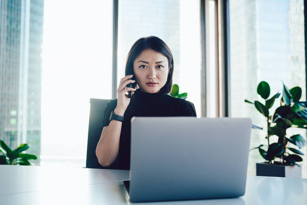 Portret azjatyckiej menedżerki siedzącej przy stole z laptopem i prowadzącej rozmowę ze smartfonem z partnerem biznesowym, chiński szef korporacyjny dzwoniący do sekretarki łączącej się z 4g na telefon komórkowy - Zdjęcie, obraz