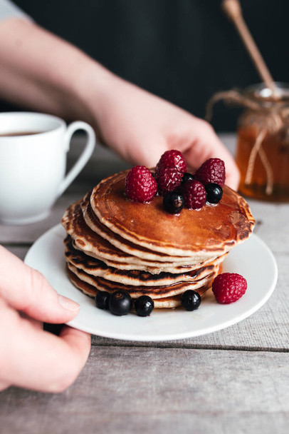 Hands hold white plate with pancakes, berries, honey, coffee cup on wooden table, jar and spoon. High quality photo - Foto, imagen