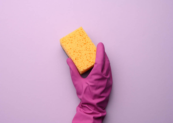 female hand in a pink rubber glove holds a yellow kitchen sponge on a purple background, close up - Photo, image