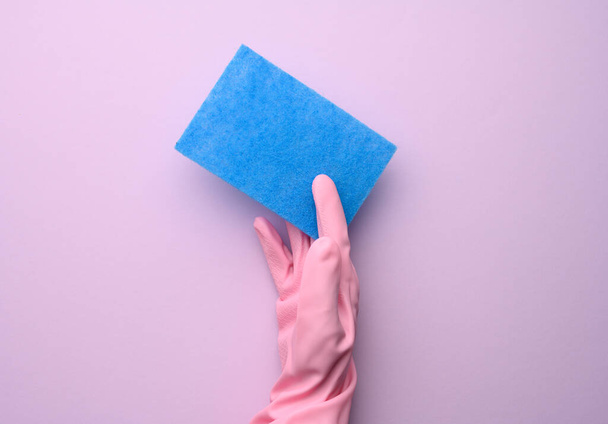 female hand in pink rubber glove holds blue kitchen sponge on purple background, close up - Zdjęcie, obraz