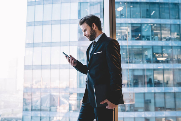 Serious confident male manager in suit holding mobile phone checking notification about income mail near panoramic windows in office, confident businessman read information from web page on cellular - Foto, Bild