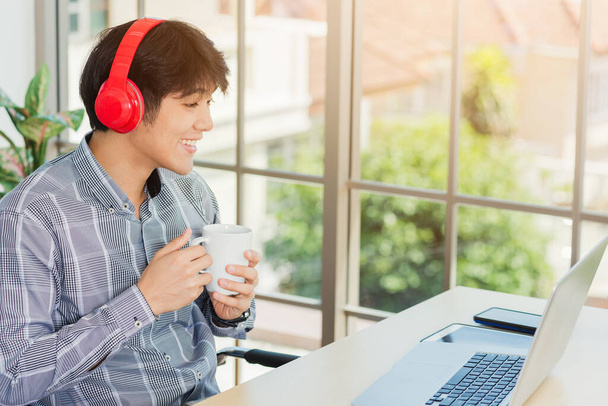 Asian young businessman happiness with red headphones sitting on desk workplace home office with a laptop computer, confident handsome man lifestyle smile relax holding a coffee cup, listens to music - Photo, image