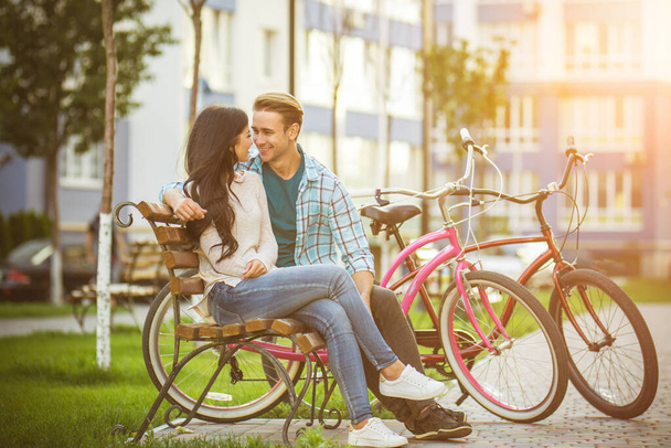 Το ευτυχισμένο ζευγάρι με ποδήλατα κάθεται σε ένα παγκάκι κοντά στο μοντέρνο σπίτι - Φωτογραφία, εικόνα