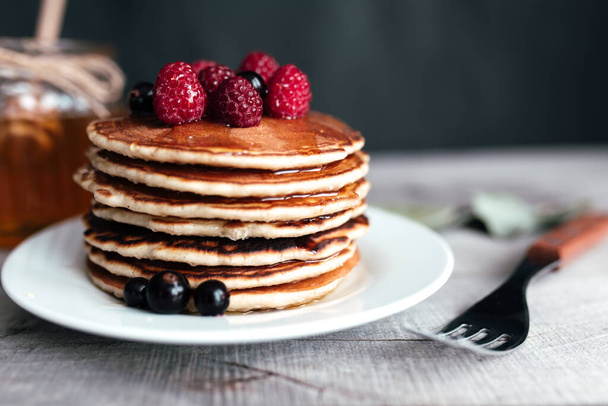 Juicy pancakes with berries and honey on a white plate, spoon, jar, wooden table. High quality photo - Foto, afbeelding