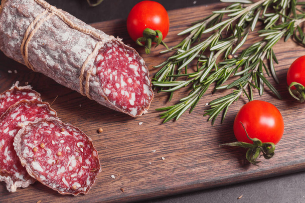 Smoked ham sausage and cutted slices with rosemary and cherry tomatoes on dark wooden board - Fotografie, Obrázek