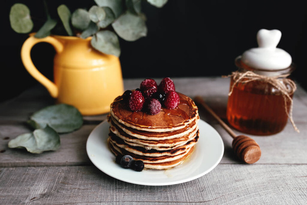 Juicy pancakes with berries and honey on a white plate, spoon, jar, wooden table, yellow vase with eucalyptus. High quality photo - Photo, Image