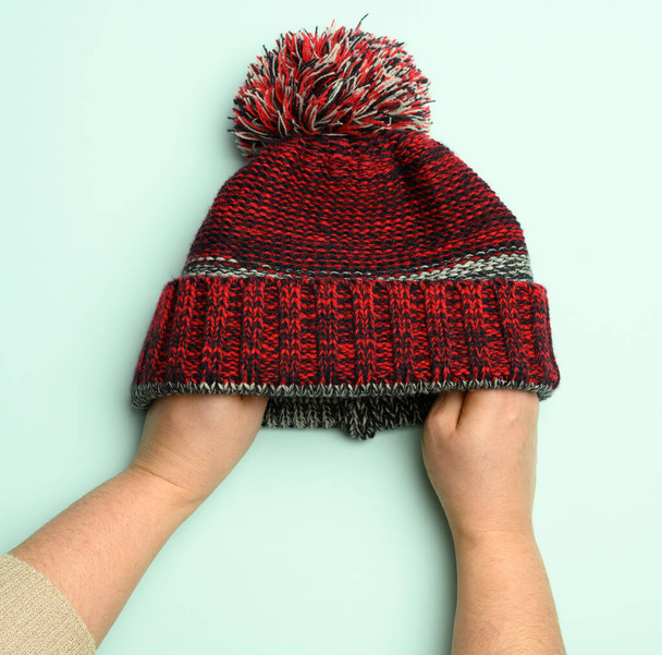 hand holding winter knitted red-black hat with pompom on a green background, comfortable clothes - Φωτογραφία, εικόνα