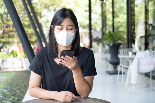 Young Asian woman wearing medical mask using smartphone at coffee shop. Asian female reading text message or social media on cell telephone during in modern cafe. New normal and social distance working. - Фото, изображение