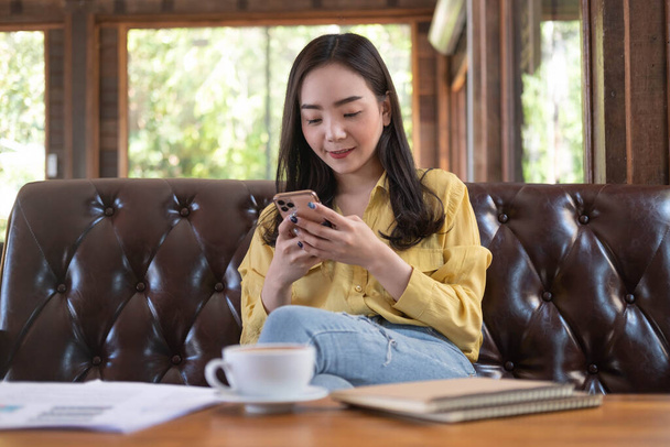 Улыбающаяся молодая азиатка пользуется смартфоном в кофейне. Азиатская женщина читает текстовые сообщения или социальные сети по мобильному телефону во время в современном кафе. Концепция системы онлайн-технологий. - Фото, изображение