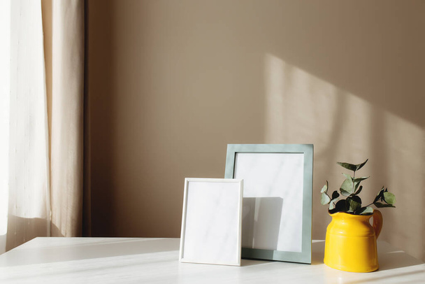 A yellow ceramic jug or vase with eucalyptus branches, empty white photo frames on the white table in the interior with beige walls near window. - Foto, afbeelding