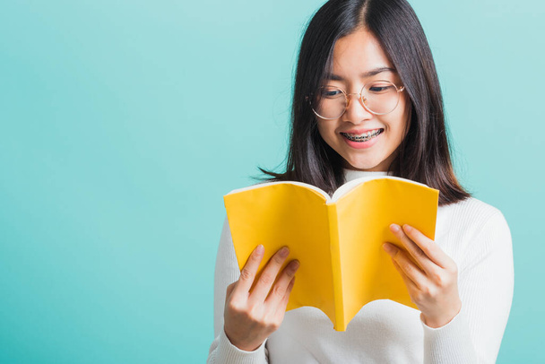 Portrait female in glasses is holding and reading a book, Young beautiful Asian woman hiding behind an open book, studio shot isolated on a blue background, Education concept - Foto, imagen