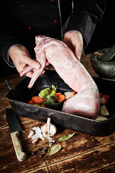 Chef preparing wild rabbit for roasting in a rustic kitchen displaying the cleaned skinned carcass to the view in a roasting pan with diced vegetables and seasoning - Zdjęcie, obraz
