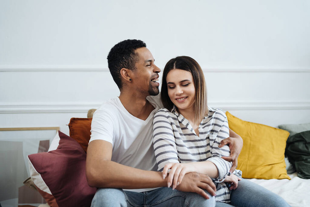 Front view of young diverse couple sitting on a couch at home - Фото, изображение