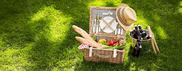 Vintage picnic hamper with vegetarian food and French baguettes adorned with an old straw hat alongside a silver cooler with bottle of beer on green grass - 写真・画像
