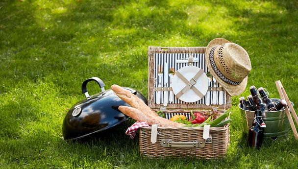 Frontal view of picnic basket and black metal barbecue next to bucket of beer bottles sitting in grass - Photo, image