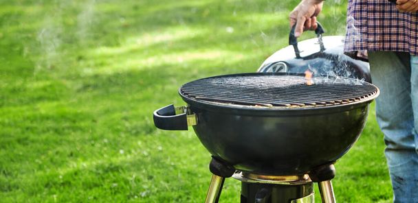 Man checking on the hot coals in a portable BBQ outdoors on a green lawn with copy space in panorama banner format - Valokuva, kuva