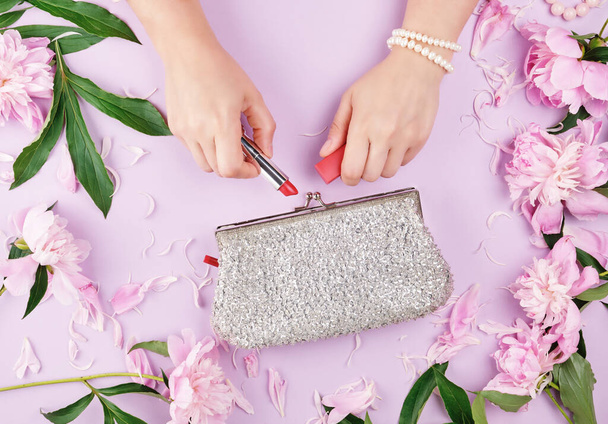 women's hands with smooth, light-skinned hold red lipstick, next to a silver clutch and a bouquet of pink peonies - Фото, зображення