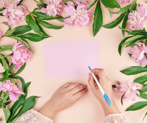 female hand holds hand a white pen over empty pink sheet of paper, next to a bouquet of blooming pink peonies, top view - Foto, afbeelding