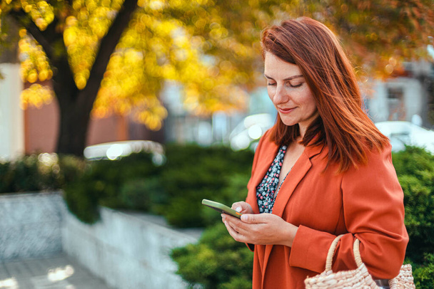 Krásná podnikatelka ve stylovém oblečení ve svých rukou používá chytrý telefon zařízení v městském parku v krásném slunečném podzimním počasí - Fotografie, Obrázek