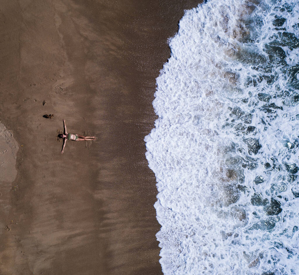 Aerial top view of woman with her hands outstretched laying on the sandy beach near the ocean. Summer holiday concept - Fotó, kép