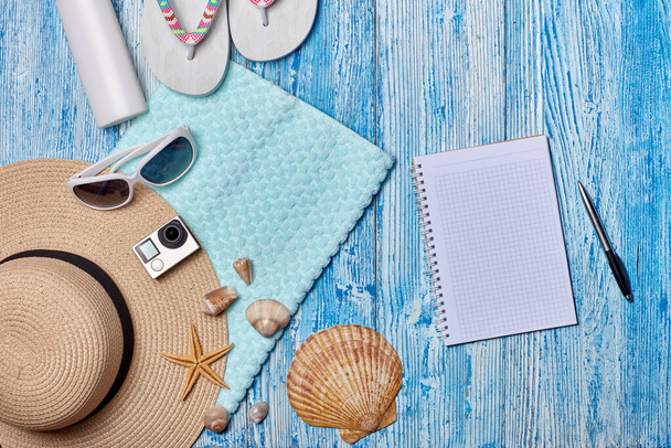 ビーチアクセサリー:フリップフロップ、ストロー帽子、サングラス、日焼け止め、タオル、青い木製の背景に空白のノートブック。コピースペース付きのトップビュー。計画休暇や旅行の概念。夏の背景 - 写真・画像