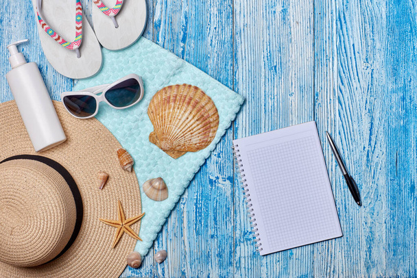 Beach accessories: flip flops, straw hat, sunglasses, sunscreen, towel, blank notebook on blue wooden background. Top view with copy space. Concept of planning vacation and travel. Summer background - 写真・画像