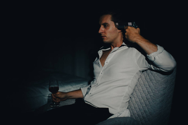 Un joven con ropa casual de negocios sentado solo en el dormitorio oscuro sosteniendo una pistola apuntando a su cabeza y copa de vino parece que va a suicidarse. Concepto de desesperanza y renunciar a las personas. - Foto, Imagen