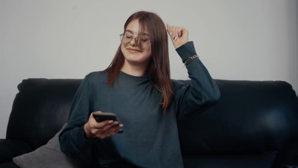 Het portret van een roodharig, sproeten meisje met bril kijkt naar het telefoonscherm en voert ontspannen danspasjes uit. Gekleed in een donkergroene blouse, zittend op een zwarte bank. Het concept van de vrouw - Video