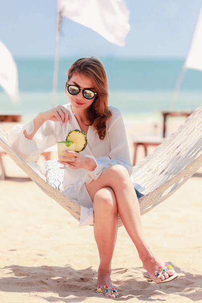 Woman wearing sun protect sunglasses drinking juice in pineapple fruit while sitting on fabric swing on white sand beach with light blue sky in background. Concept of outdoor nature travel at weekend. - Photo, Image