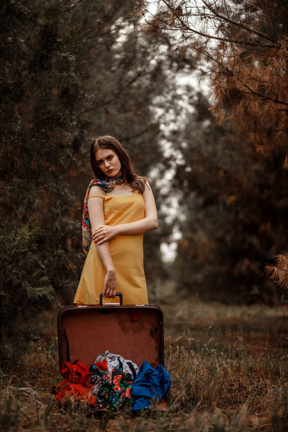 portrait of a young beautiful girl in a yellow dress standing in the rain with an open vintage suitcase with fallen clothes - Valokuva, kuva