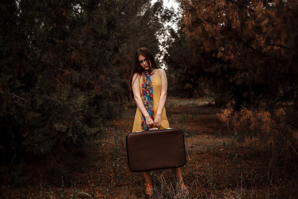 portrait of a young beautiful girl in a yellow dress with a colorful scarf standing in the woods with a vintage suitcase in hand - Fotoğraf, Görsel