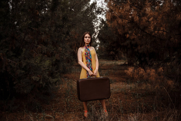 portrait of a young beautiful girl in a yellow dress with a colorful scarf standing in the woods with a vintage suitcase in hand - Fotoğraf, Görsel