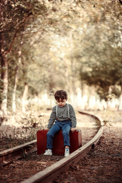 little cute boy in vintage clothes standing on an abandoned railroad with a vintage suitcase in his hands - Foto, imagen