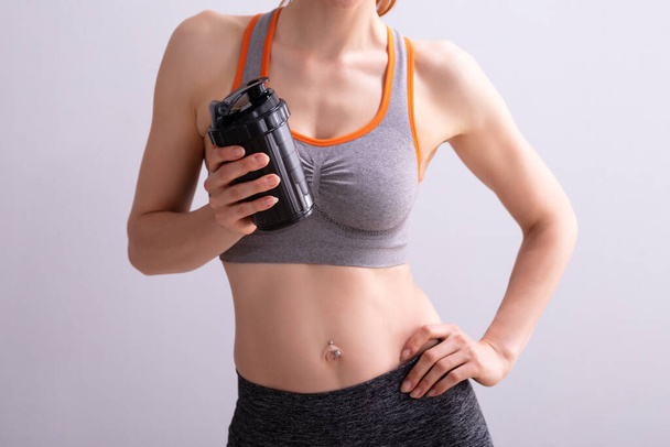 Portrait Of Smiling Fitness Woman Holding Protein Shaker Bottle Against Gray Background - 写真・画像