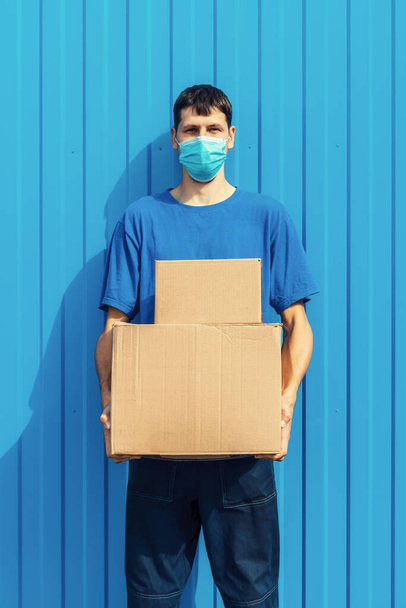 a young Caucasian courier volunteer in a medical mask holds cardboard boxes in his hands. - Foto, imagen