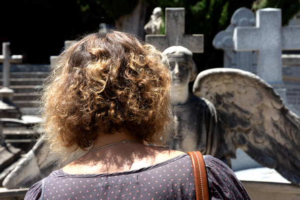 Lähikuva tuntemattoman naisen selästä, joka vierailee haudoissa suojelusenkelin epätarkan patsaan edessä. Kiharat vaaleat hiukset ja osa naisen hartioista näkyvät. - Valokuva, kuva