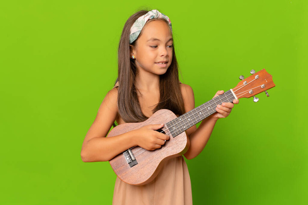 piękne uśmiechnięta dziewczyna uczy się grać ukulele izolowane na zielonym tle. - Zdjęcie, obraz