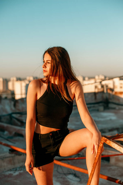 portrait of a young brunette girl in black sneakers,black shorts and a black short top standing on the roof and enjoying the sunset - Фото, изображение