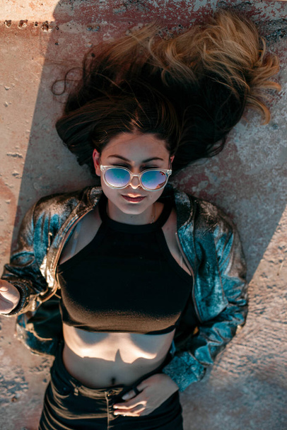 portrait of a young brunette girl with a beautiful body in black clothes and a silver jacket lying on the roof and enjoying the sunset - Foto, imagen