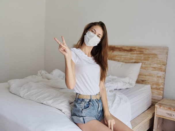 Tıbbi maskeli güzel bir kadın parlak bir odada bir yatakta oturuyor ve el hareketi yapıyor. - Fotoğraf, Görsel