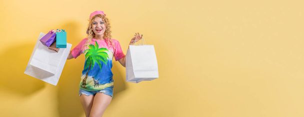 Boldog fiatal bolti lány fényes alkalmi ruhák áll a sárga háttérben. Kézben tartja a táskákat a vásárlásokkal. A kamerába néz.. - Fotó, kép