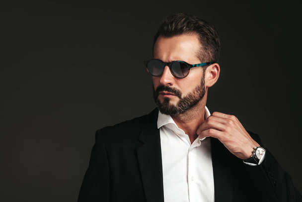 Closeup portrait of handsome confident stylish hipster lambersexual model. Sexy modern man dressed in elegant black suit. Fashion male posing in studio on dark background. In sunglasses - Photo, Image