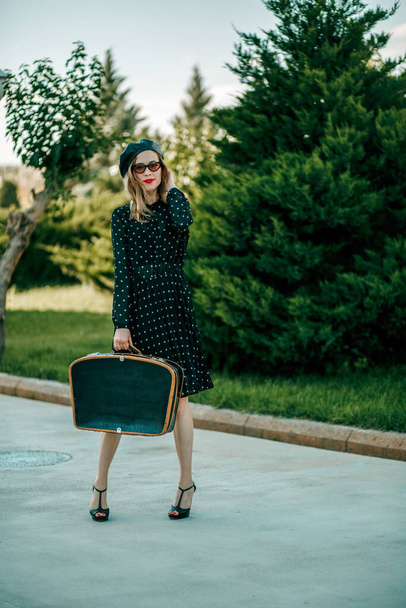 young woman in vintage black polka dot dress with retro suitcase in hand posing outside - Фото, изображение
