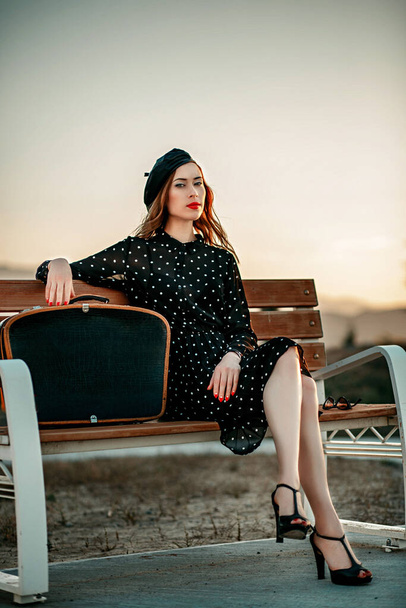 young woman in vintage black polka dot dress with retro suitcase in hand posing outside - 写真・画像