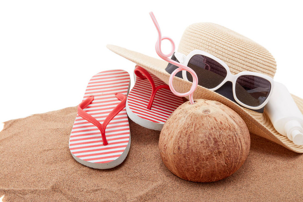 Strand accessoires: teenslippers, strohoed, zonnebril, zonnebrandcrème en kokosnoot, liggend op het zand op witte achtergrond. Concept van de zee vakantie en reizen. Zomer achtergrond, Bescherming tegen uv - Foto, afbeelding