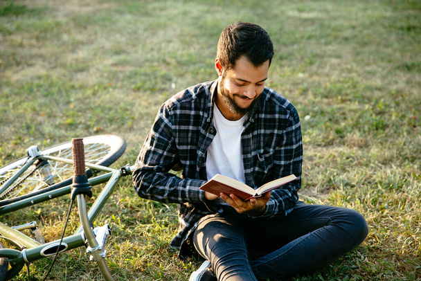 Attractive smiling bearded man reading interesting book, spending time with pleasure while sitting on the grass near the bike. Leisure concept. - Foto, Bild