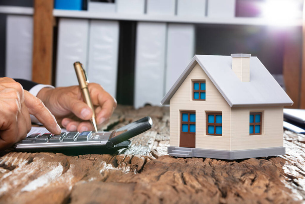 Close-up Of House Model In Front Of Businessperson's Hand Using Calculator - Foto, Imagem