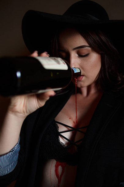 beautiful young girl in black underwear and black pantsuit in the entrance drinking wine or drink from a bottle. - Photo, image
